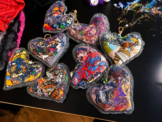 Seven colorful heart-shaped fabric keychains with mixed-media artwork, displayed on a black surface.