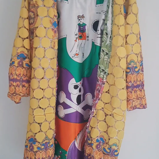 Close-up view of a vibrant, intricately designed fabric detail from an upcycled vintage kimono-style coat. Features bold yellow lace patterns, colorful floral embroidery, and a mix of rich, festive prints inspired by Canarian carnival aesthetics. A striking combination of textures and colors for a unique, eye-catching garment.